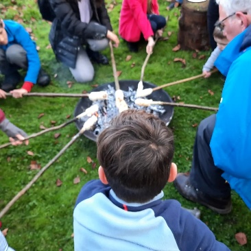 A young child prepares to start a campfire while others gather around in a wooded outdoor setting.AI-generated content may be incorrect.